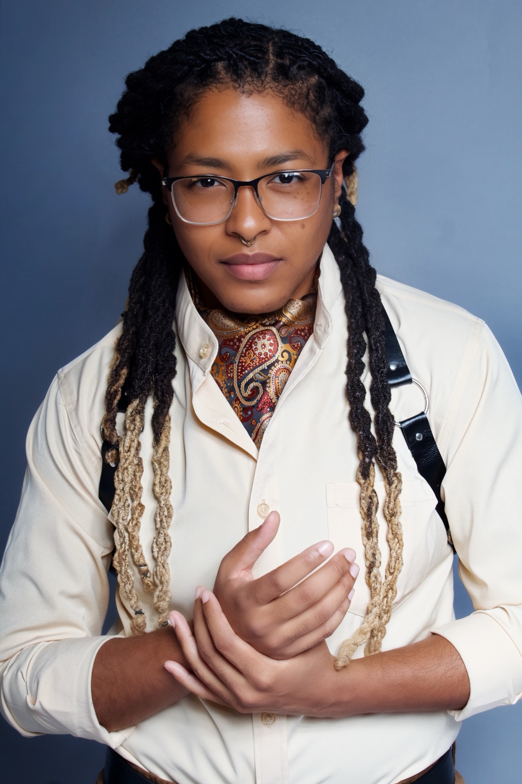 Person with glasses and long locs in a cream shirt against a blue-gray backdrop