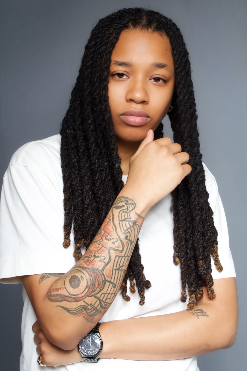 Creative professional with long locs and tattooed arms in a white shirt against a gray backdrop